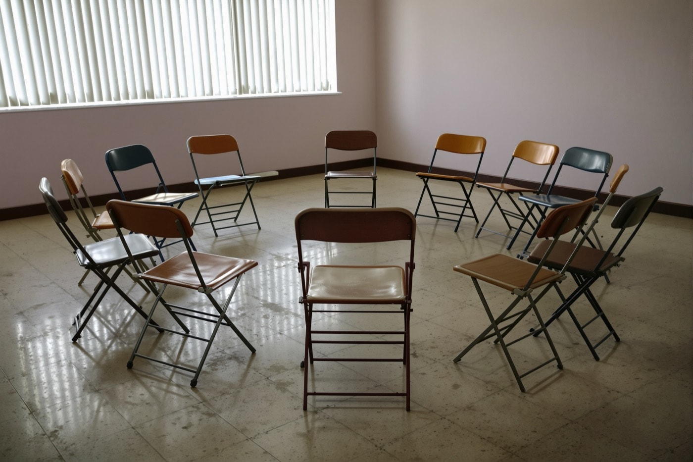 An empty circle of folding chairs in a plain community meeting room with morning light through blinds